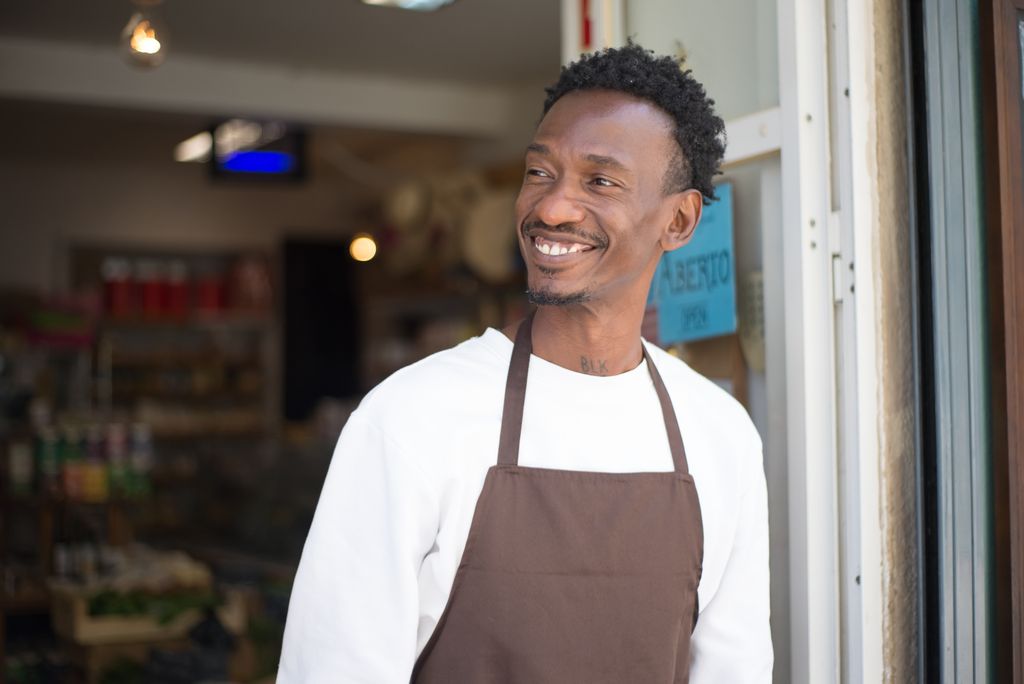 Photo by Kampus Production Smiling store owner in brown apron standing at entrance, welcoming customers.