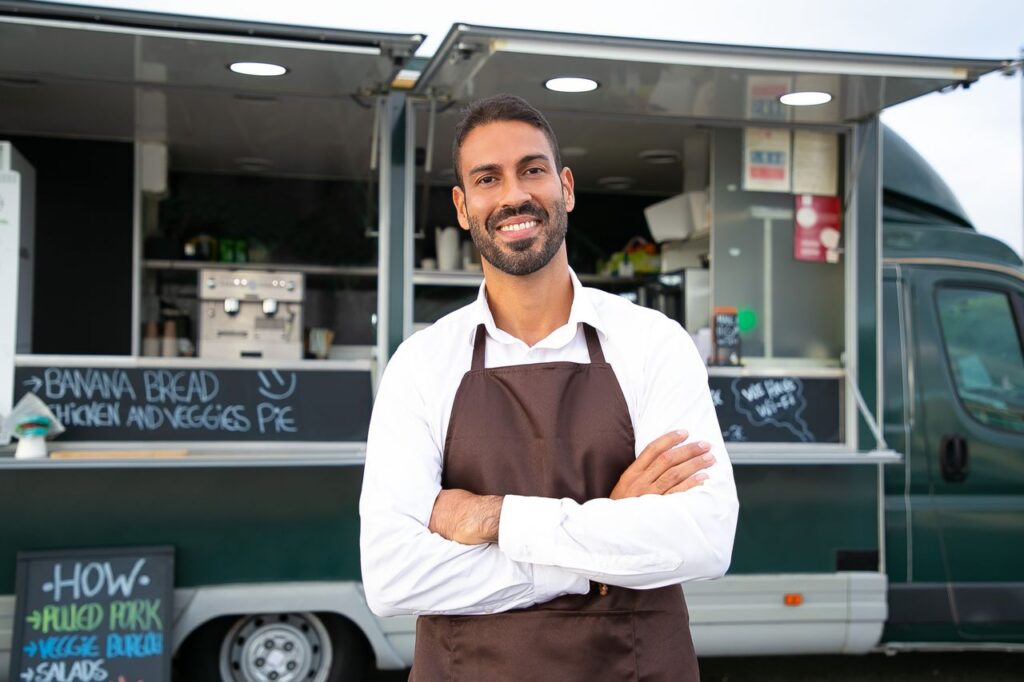 pexels-photo-5920774-5920774 Smiling young ethnic male waiter in apron standing with arms folded near modern food truck and looking at camera contentedly